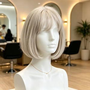 Könnte beinhalten: Ein weißer Schaufensterpuppenkopf mit einer schulterlangen, platinblonden Bob-Perücke mit weichem Pony. Das Haar ist glatt und mit dezenten Stufen gestylt. Eine zarte goldene Kette mit einem kleinen Anhänger liegt am Hals der Schaufensterpuppe.