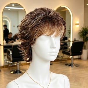 May include: A short, layered wig with a shaggy, textured cut featuring a blend of warm brown and light blonde highlights. The hair is styled with wispy bangs that frame the forehead, resting on a smooth, white, featureless mannequin head.