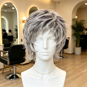 May include: A mannequin head wearing a short, layered wig with a shaggy, textured cut. The hair is a blend of silver, white, and light gray tones, styled with wispy bangs and flipped ends that frame the face. The mannequin has a smooth white finish.