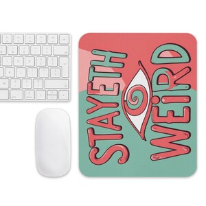 May include: A white wireless keyboard and mouse set next to a rectangular mousepad. The mousepad has a red and teal background with the words "STAYETH WEIRD" and an eye graphic.