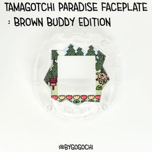 Pode incluir: Uma placa frontal transparente do Tamagotchi Paradise com um design Brown Buddy Edition. A placa frontal apresenta uma paisagem colorida com árvores, um urso e flores. O texto "TAMAGOTCHI PARADISE FACEPLATE : BROWN BUDDY EDITION" está no topo.