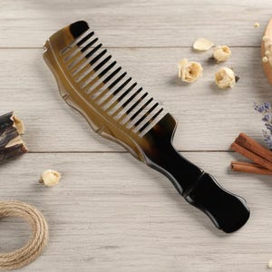 May include: A handcrafted comb with a dark brown handle and a light brown comb section. The comb features evenly spaced black teeth. The handle has a curved design for comfortable grip. The comb is made of natural materials.