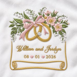 Pode incluir: Um design bordado em tecido branco apresentando dois anéis dourados entrelaçados com uma pequena pedra branca, acentuado por flores cor-de-rosa, folhas verdes e lírios do vale. O texto diz William and Jordyn com a data 08 01 2026.