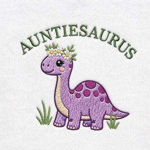 May include: Embroidered design of a purple dinosaur with a daisy crown and pink spots. The text "AUNTIESAURUS" arches above the dinosaur in green. Green grass is at the bottom of the image, all on a white background.