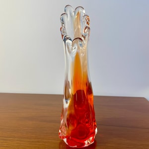 May include: A tall, slender glass vase featuring a clear exterior and a vibrant, swirling interior pattern of deep red and bright orange hues. The rim is uniquely shaped with a scalloped, ruffled edge, and the base has a textured, organic form.