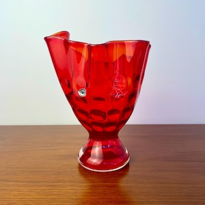 May include: A vibrant red glass vase featuring a textured, dimpled surface and a flared, asymmetrical rim. The vase stands on a circular pedestal base and includes a small, circular silver-coloured sticker with the text FANY on the side.