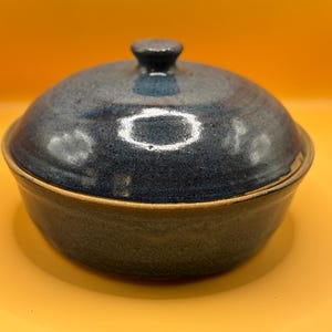 Puede incluir: Un cuenco de cerámica con tapa. El cuenco y la tapa son de un azul oscuro moteado con un borde marrón. La tapa tiene un pequeño pomo redondeado. El cuenco está sobre una superficie amarilla.