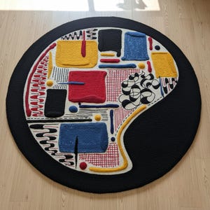 May include: A round rug with a black border and a colorful abstract design. The interior features squares and lines in yellow, red, blue, and black, with a white background. The rug has a textured, tufted appearance.