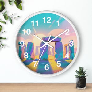 May include: White-framed wall clock featuring a colorful illustration of Stonehenge. The clock face has a teal and pink sky with the monument in shades of purple and blue. White hands and numerals complete the design, with a small plant in a pot on the right.