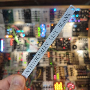 May include: A white label with the text "INTENTIONALLY LEFT BLANK" is held in front of a complex electronic circuit board. The background is filled with various modules, wires, and colourful lights.