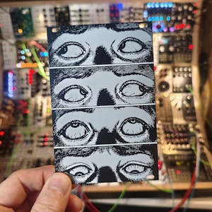 May include: A monochrome panel displaying four sets of stylized eyes with an intense gaze. The artwork is held in front of a complex electronic device with various components and wires, suggesting a connection to audio or electronic music.