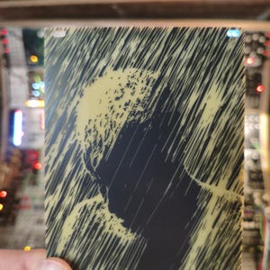 Peut inclure: Une carte rectangulaire avec une silhouette noire d'une tête et des épaules, sur un fond jaune de lignes diagonales. La carte est tenue dans une main, avec des composants électroniques visibles en arrière-plan.
