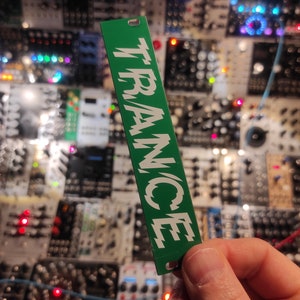 Puede incluir: Una etiqueta rectangular verde con la palabra "TRANCE" escrita en blanco, con letras estilizadas.
