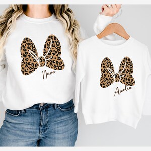 Op de afbeelding: Witte sweatshirts met een luipaardprint strik ontwerp. E&eacute;n sweatshirt heeft de naam "Nana" onder de strik gedrukt, en de andere heeft "Amelia". De sweatshirts worden getoond op een persoon en een hanger.
