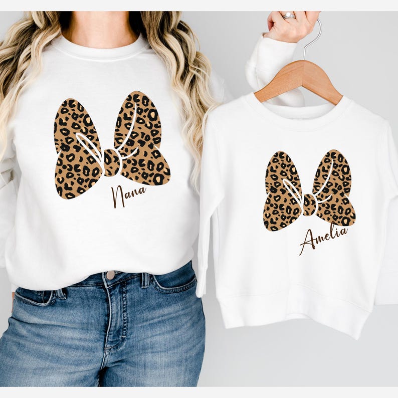 Op de afbeelding: Witte sweatshirts met een luipaardprint strik ontwerp. E&eacute;n sweatshirt heeft de naam "Nana" onder de strik gedrukt, en de andere heeft "Amelia". De sweatshirts worden getoond op een persoon en een hanger.