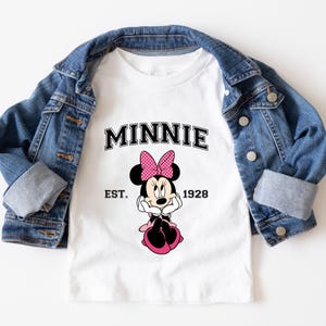 May include: White t-shirt featuring the word "MINNIE" in black collegiate lettering, with a pink polka-dot bow and Minnie Mouse graphic. The text "EST. 1928" is below the graphic. A blue denim jacket is draped over the shirt.