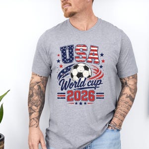 May include: A heather gray t-shirt featuring a soccer ball and the text "USA World Cup 2026" in red, white, and blue. The design includes stars and stripes, celebrating the upcoming soccer tournament.