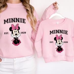 May include: Pink sweatshirts featuring a Minnie Mouse graphic. The design includes the word "MINNIE" above an image of Minnie Mouse, with "EST. 1928" below. The Minnie Mouse graphic has a pink bow and black ears.