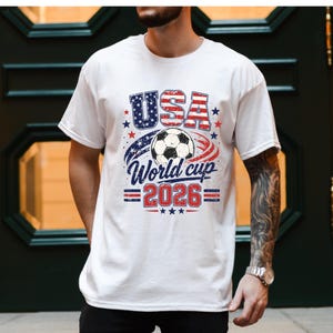 May include: White t-shirt with a graphic design featuring the word "USA" in red and blue, a soccer ball, and the text "World Cup 2026". The design incorporates stars and stripes, celebrating the upcoming soccer tournament.