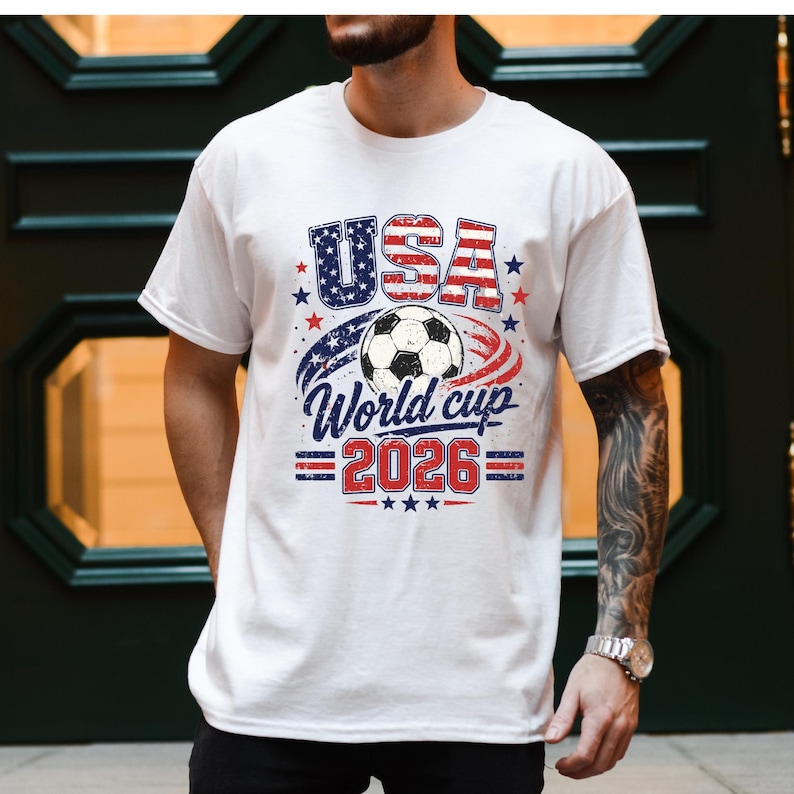 May include: White t-shirt with a graphic design featuring the word "USA" in red and blue, a soccer ball, and the text "World Cup 2026". The design incorporates stars and stripes, celebrating the upcoming soccer tournament.