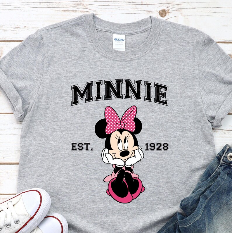 May include: A gray t-shirt featuring the word "MINNIE" in black block letters above a cartoon image of Minnie Mouse. The image includes a pink bow with white polka dots, and the text "EST. 1928" below the image. White sneakers and denim jeans are visible.
