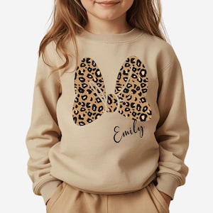 Op de afbeelding: Beige sweatshirt met een luipaardprint strik ontwerp. De strik is in bruin en zwart. De naam "Emily" staat eronder in cursief geschreven. De sweatshirt heeft lange mouwen en een ronde hals.