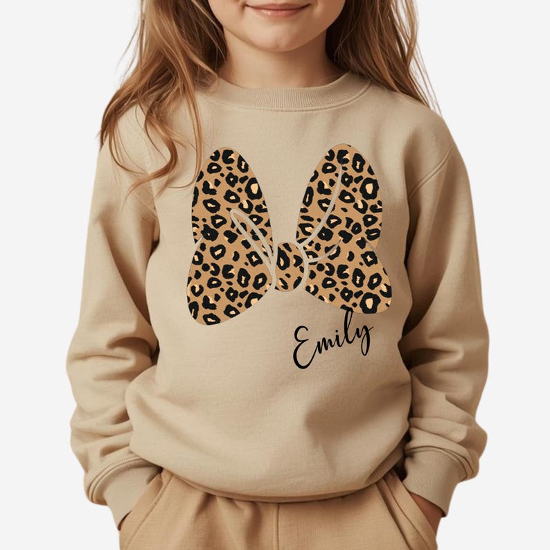 Op de afbeelding: Beige sweatshirt met een luipaardprint strik ontwerp. De strik is in bruin en zwart. De naam "Emily" staat eronder in cursief geschreven. De sweatshirt heeft lange mouwen en een ronde hals.