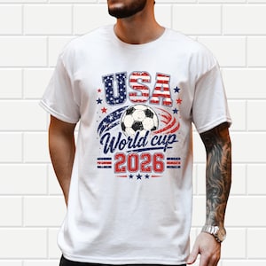 May include: White t-shirt with a graphic design for the 2026 World Cup. The design features the word "USA" in red and blue, a soccer ball, and the text "World Cup 2026". The shirt has a distressed, vintage look.