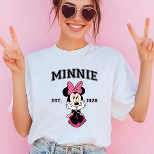 May include: White t-shirt featuring a Minnie Mouse graphic. The design includes the name "MINNIE" in a black, block font above an illustration of Minnie Mouse wearing a pink bow. The text "EST. 1928" is below the character.