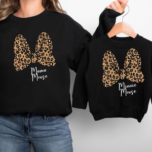 Op de afbeelding: Zwarte sweatshirts met een luipaardprint strik ontwerp. E&eacute;n sweatshirt zegt "Mama Mouse" en de andere zegt "Minnie Mouse" in een scriptlettertype. De sweatshirts zijn een bijpassende set.