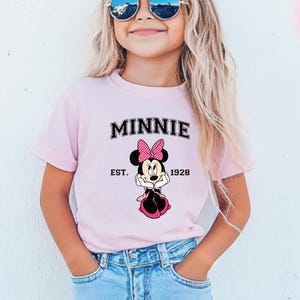 May include: A light pink t-shirt featuring the word "MINNIE" in black block letters above a Minnie Mouse graphic. The graphic includes a pink polka dot bow and the text "EST. 1928". The shirt is worn by a person wearing sunglasses.