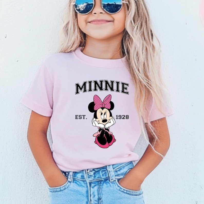 May include: A light pink t-shirt featuring the word "MINNIE" in black block letters above a Minnie Mouse graphic. The graphic includes a pink polka dot bow and the text "EST. 1928". The shirt is worn by a person wearing sunglasses.