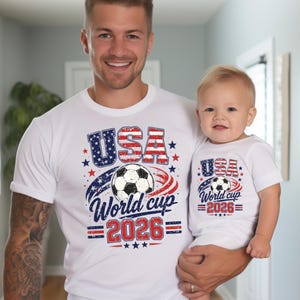 May include: White t-shirts with a patriotic design. The shirts feature the text "USA World Cup 2026" with a soccer ball graphic, stars, and red and blue accents. One shirt is adult-sized, the other is for a baby.