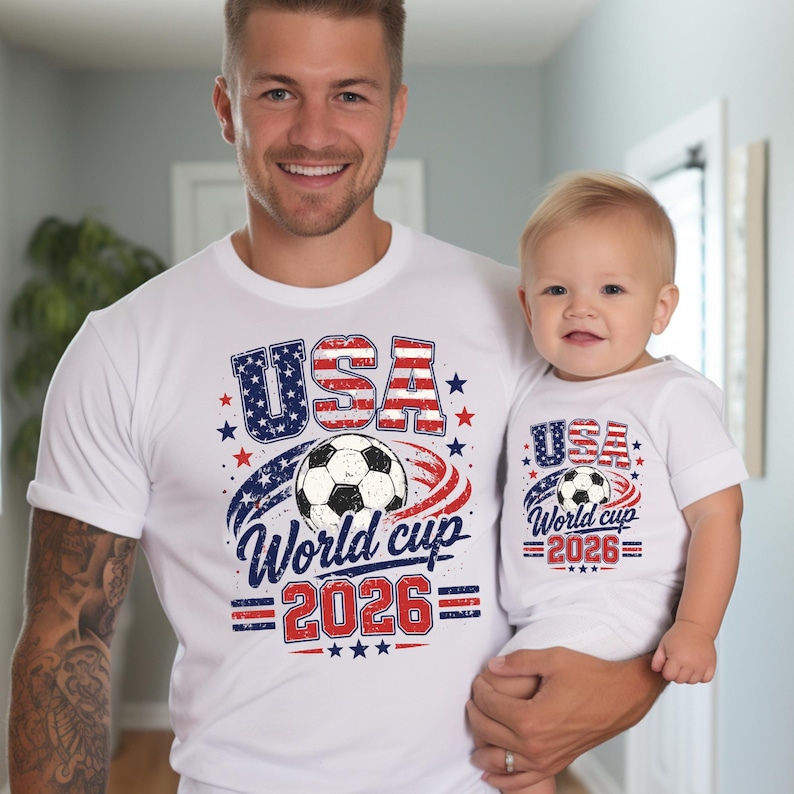 May include: White t-shirts with a patriotic design. The shirts feature the text "USA World Cup 2026" with a soccer ball graphic, stars, and red and blue accents. One shirt is adult-sized, the other is for a baby.
