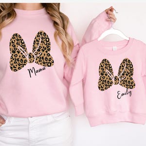 Op de afbeelding: Roze sweatshirts met bijpassende luipaardprint strik ontwerpen. E&eacute;n sweatshirt zegt "Mama" en de andere "Emily". De sweatshirts hangen aan een witte hanger.