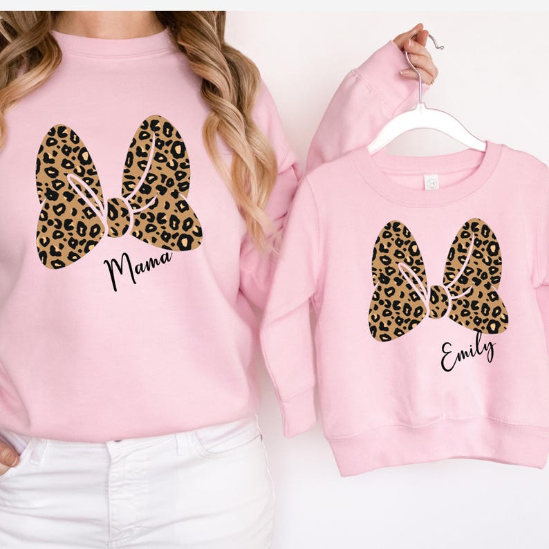 Op de afbeelding: Roze sweatshirts met bijpassende luipaardprint strik ontwerpen. E&eacute;n sweatshirt zegt "Mama" en de andere "Emily". De sweatshirts hangen aan een witte hanger.