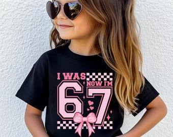 May include: A black t-shirt with the text "I WAS... NOW I'M 67" in pink and white. The design includes a bow, hearts, and checkered patterns. The shirt is worn by a young person wearing heart-shaped sunglasses.