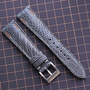 Puede incluir: Correa de reloj de dos piezas con un patrón gris texturizado. La correa presenta costuras blancas y costuras de acento naranja cerca de las asas. Una hebilla de color plateado está unida a un extremo. La correa del reloj se muestra sobre un fondo de cuadrícula.