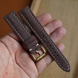 May include: A dark brown suede watch strap featuring white contrast stitching along the edges. The strap includes a polished gold-tone metal buckle and a matching leather keeper loop, designed for secure attachment to a watch case.