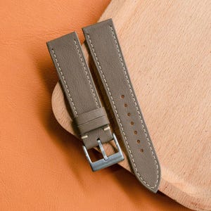 May include: Two taupe leather watch straps with white stitching and a silver buckle. The straps are displayed on a wooden tray and an orange leather surface.