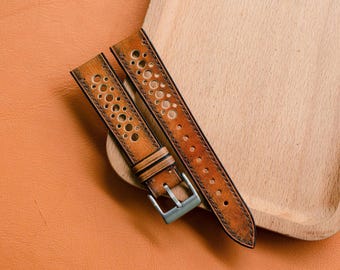 Puede incluir: Dos correas de reloj de cuero marrón con un acabado envejecido y perforaciones circulares. Las correas tienen una hebilla plateada y se muestran sobre una bandeja de madera y una superficie de cuero naranja.