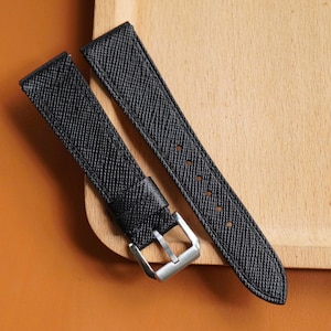 Puede incluir: Dos correas de reloj negras con superficie texturizada y hebilla plateada. Las correas están colocadas sobre una superficie de madera marrón claro y un fondo naranja.