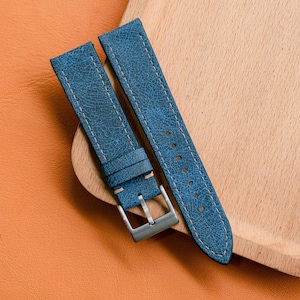 May include: Two blue leather watch straps with white stitching and a silver buckle. The straps are placed on a wooden tray, set against an orange leather surface. The leather has a distressed texture.