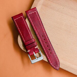 Peut inclure: Bracelet de montre en deux parties, rouge foncé, avec coutures blanches et boucle argentée. Le bracelet en cuir est présenté sur une surface en bois et un fond beige.