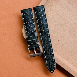 May include: Black leather watch straps with white stitching and a perforated design. The straps have a silver-coloured buckle and are displayed on a wooden surface.