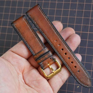 Puede incluir: Una correa de reloj de cuero marrón de dos piezas con acabado texturizado, bordes oscuros y una hebilla de metal pulido en tono dorado. La correa incluye una trabilla de cuero fija y una serie de orificios perforados para su ajuste.