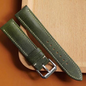 Op de afbeelding: Groene leren horlogeband met witte stiksels en een zilveren gesp. De band wordt gepresenteerd op een houten oppervlak en een bruine achtergrond. De horlogeband is ongeveer 20 cm lang.