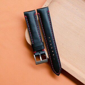 Könnte beinhalten: Schwarze Leder-Armbanduhren mit roten Nähten. Die Armbänder haben eine silberfarbene Schnalle und werden auf einem Holztablett präsentiert. Die Armbänder sind neu und unbenutzt.