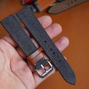 Può includere: Primo piano di un cinturino per orologio con una trama in tela grigio scuro e dettagli in pelle marrone. Il cinturino ha una fibbia argentata ed è tenuto in mano da una persona. Altri cinturini per orologi sono visibili sullo sfondo.