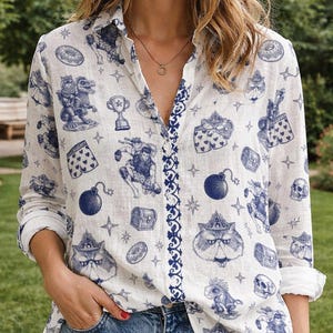 Puede incluir: Camisa blanca con botones y estampado azul caprichoso. El estampado presenta varios personajes de dibujos animados, bombas, cofres del tesoro y palos de cartas. La camisa tiene mangas largas y un ribete azul decorativo a lo largo de la tapeta.